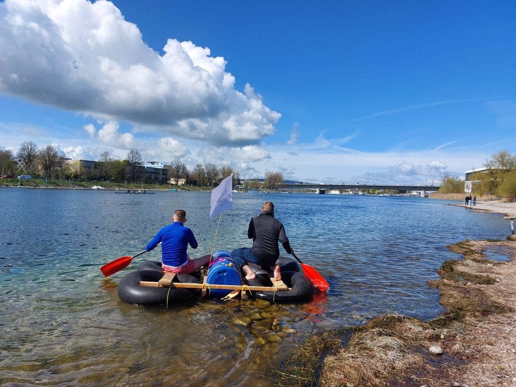 Zwei Teilnehmer paddeln mit ihrem selbstgebauten Floß und eigener Flagge auf den Bodensee